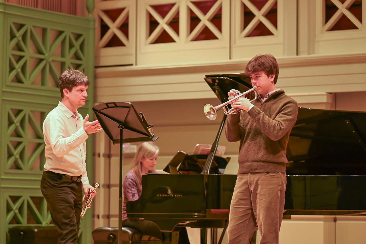 A male professor leading a male student in a trumpet performance.